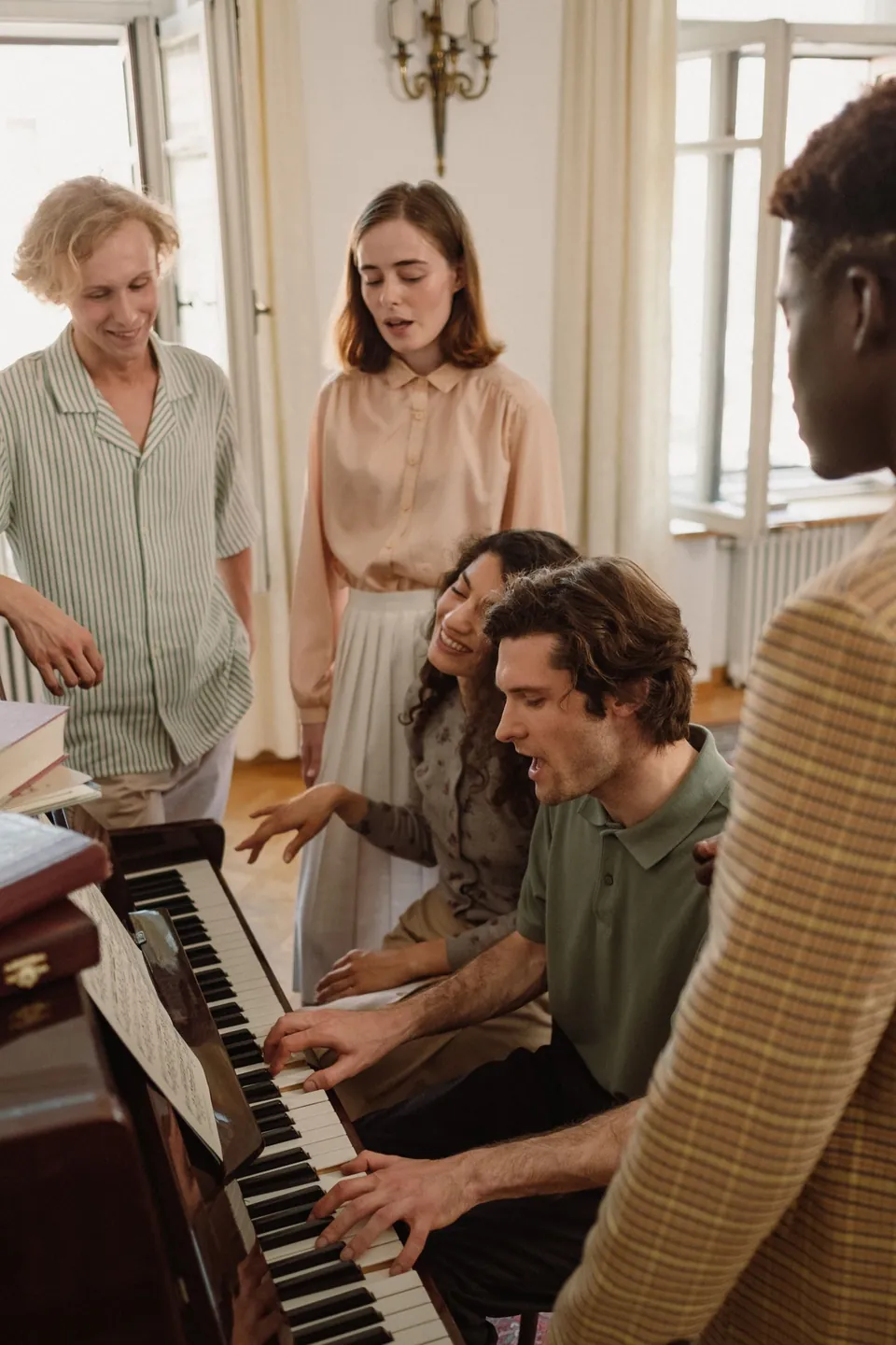 A group of adults singing together around a person playing piano indoors